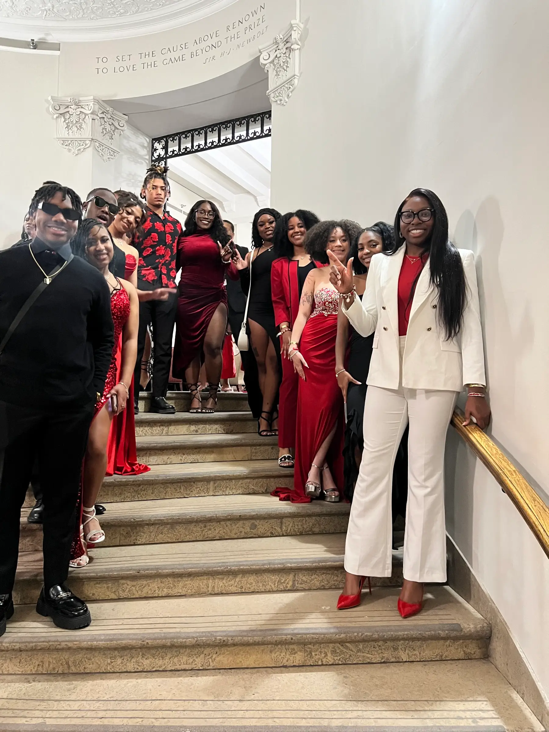 About fourteen students in red, black and white formalwear posing on a stone staircase under a carved inscription
