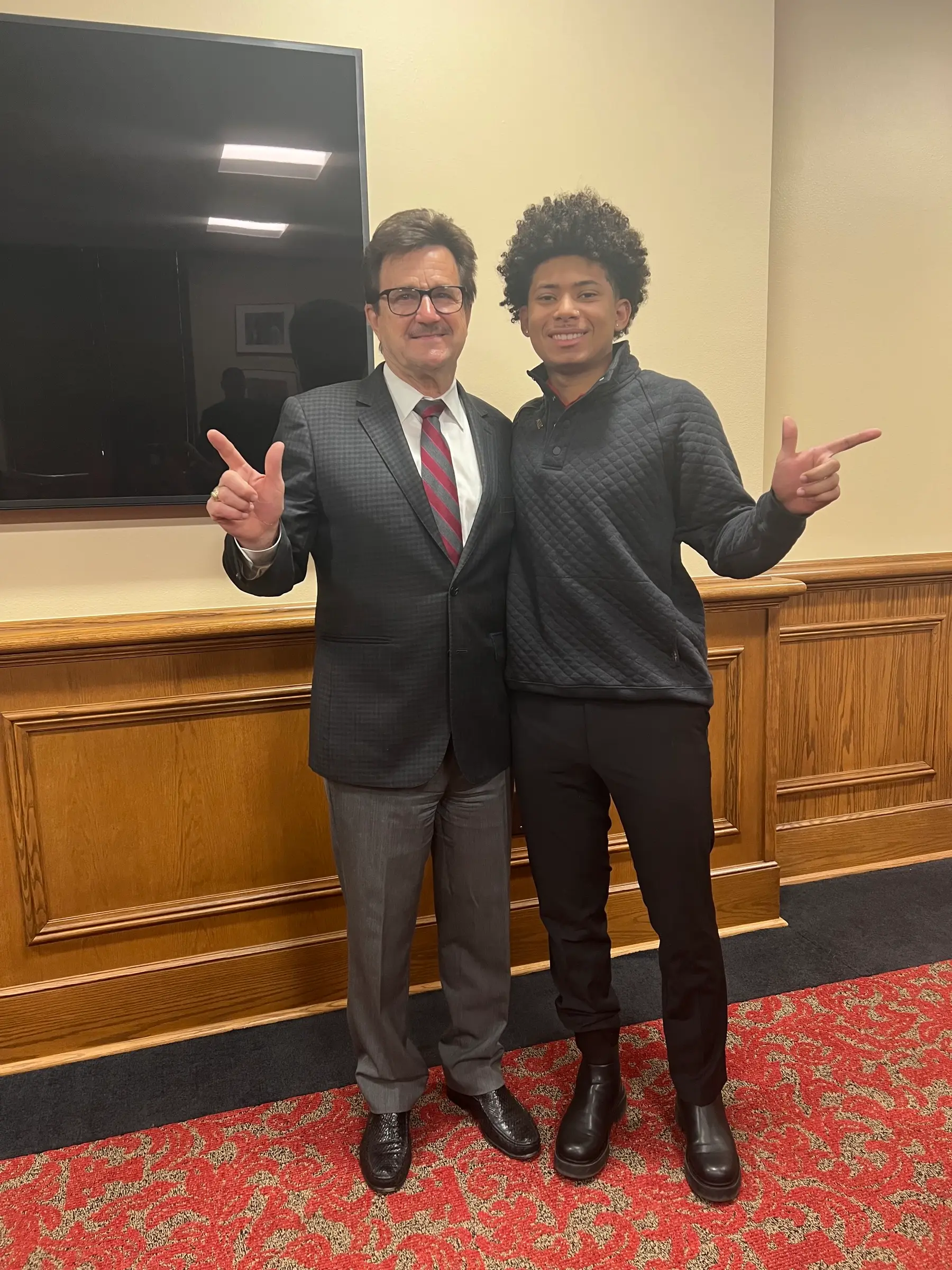 Two men giving the Guns Up hand sign in a wood-paneled room with red patterned carpet