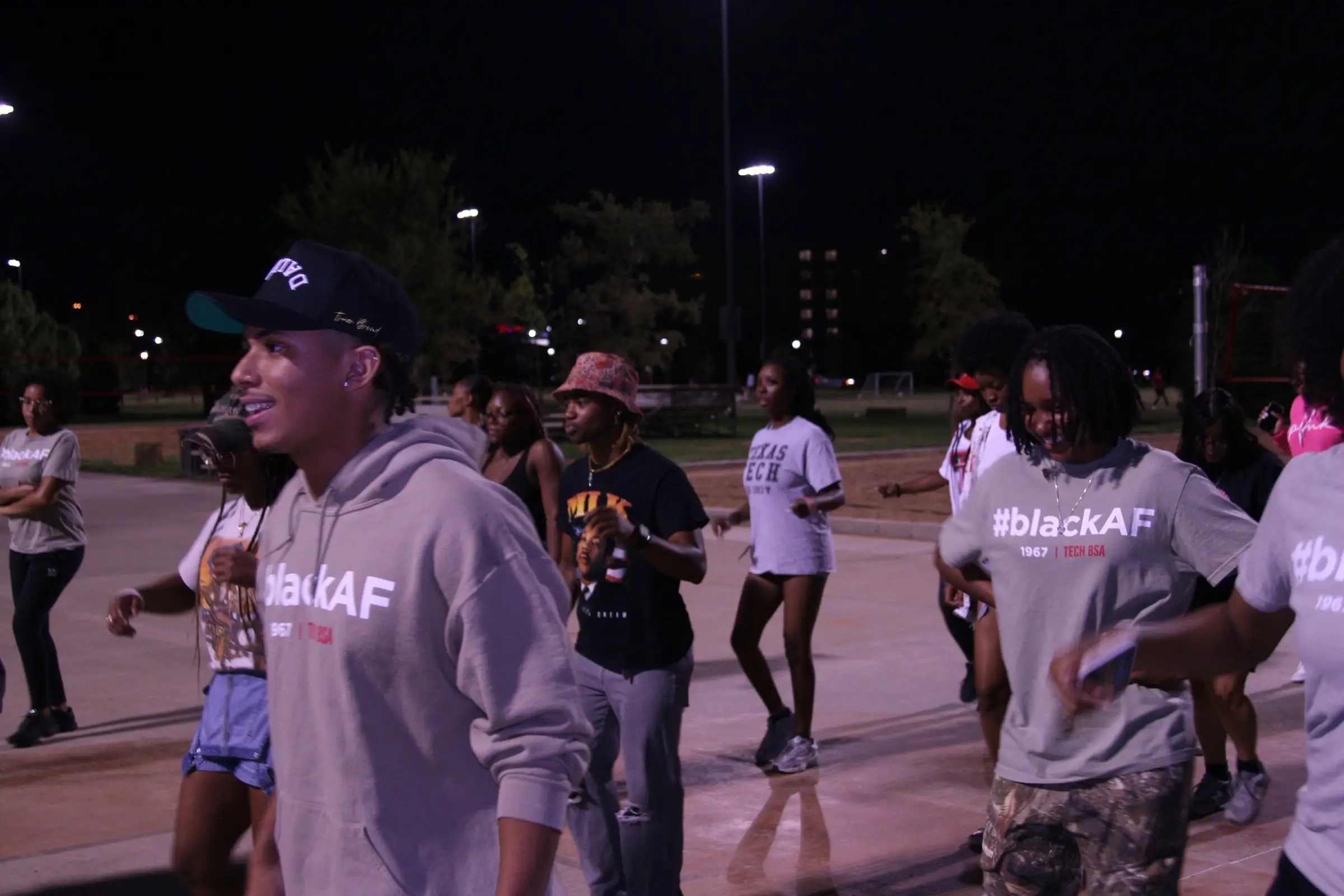 Students line dancing on a lit outdoor court at night, several wearing grey #blackAF 1967 Tech BSA shirts