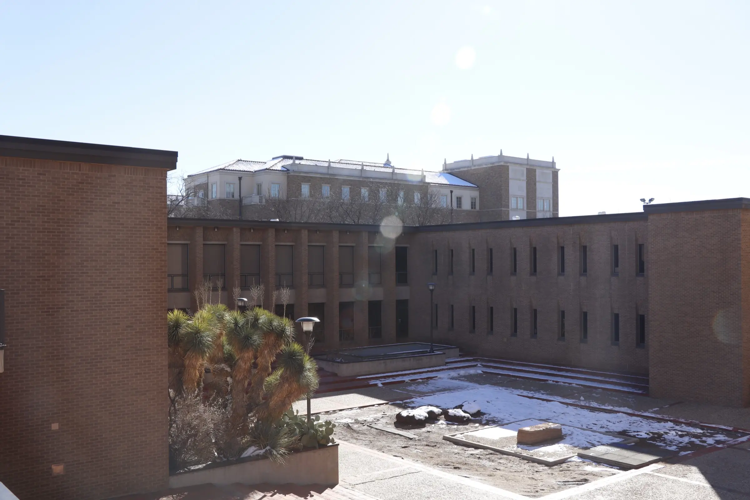 Brick campus courtyard with patchy snow and a yucca, tiled-roof building behind under a pale sky with lens flare