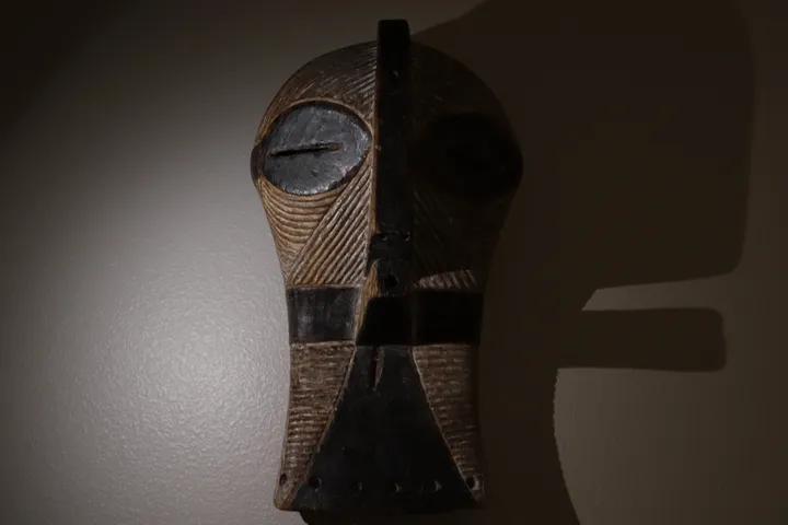 Carved wooden mask with striated grooves and dark round eyes, side-lit against a textured wall with a hard shadow
