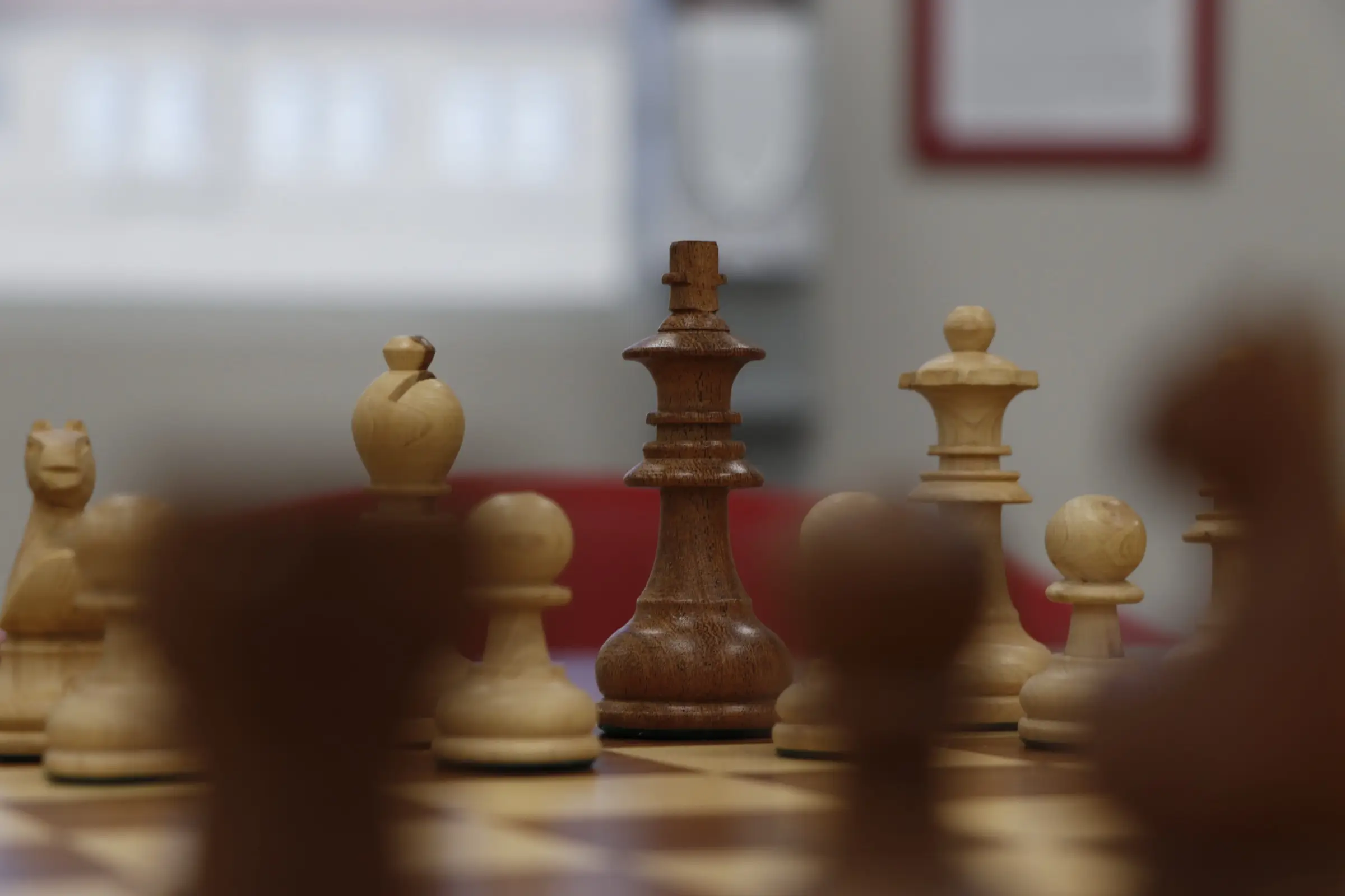 Close-up of a dark wooden chess king in focus among light pieces, blurred foreground pieces and red chair behind