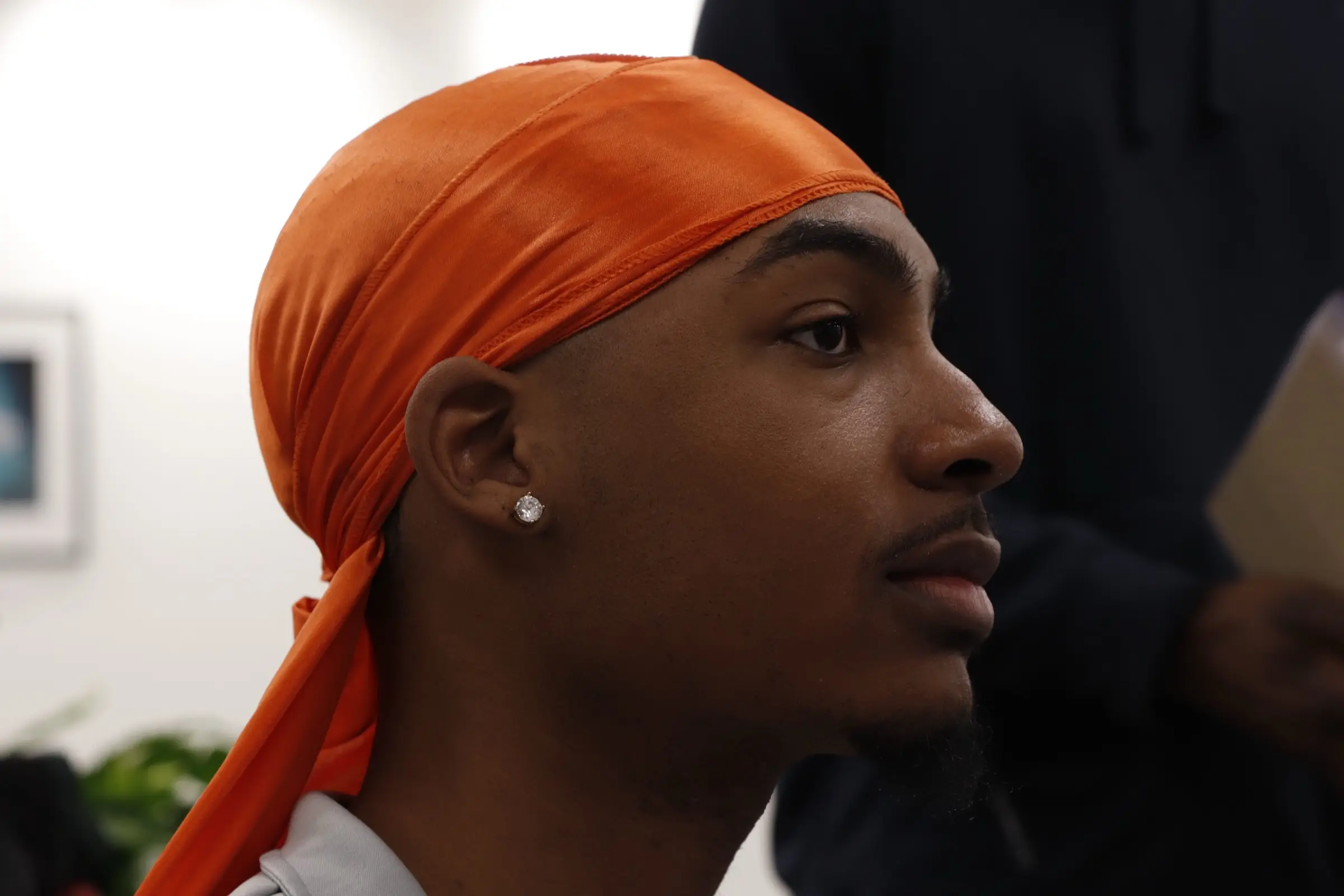Side-profile portrait of a man in an orange satin durag with a stud earring, bright blurred background