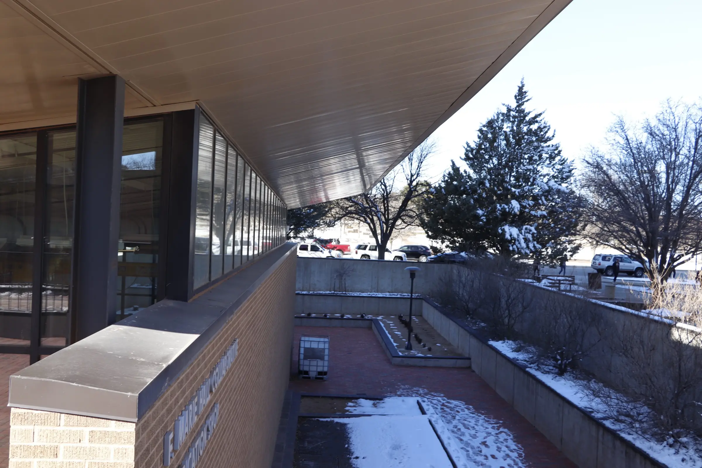 Cantilevered roof over a brick wall and window band beside a sunken snowy courtyard with evergreen trees