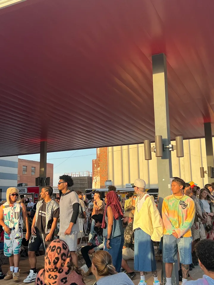 Streetwear models lined up under a red canopy at golden hour, audience seated, food trucks behind