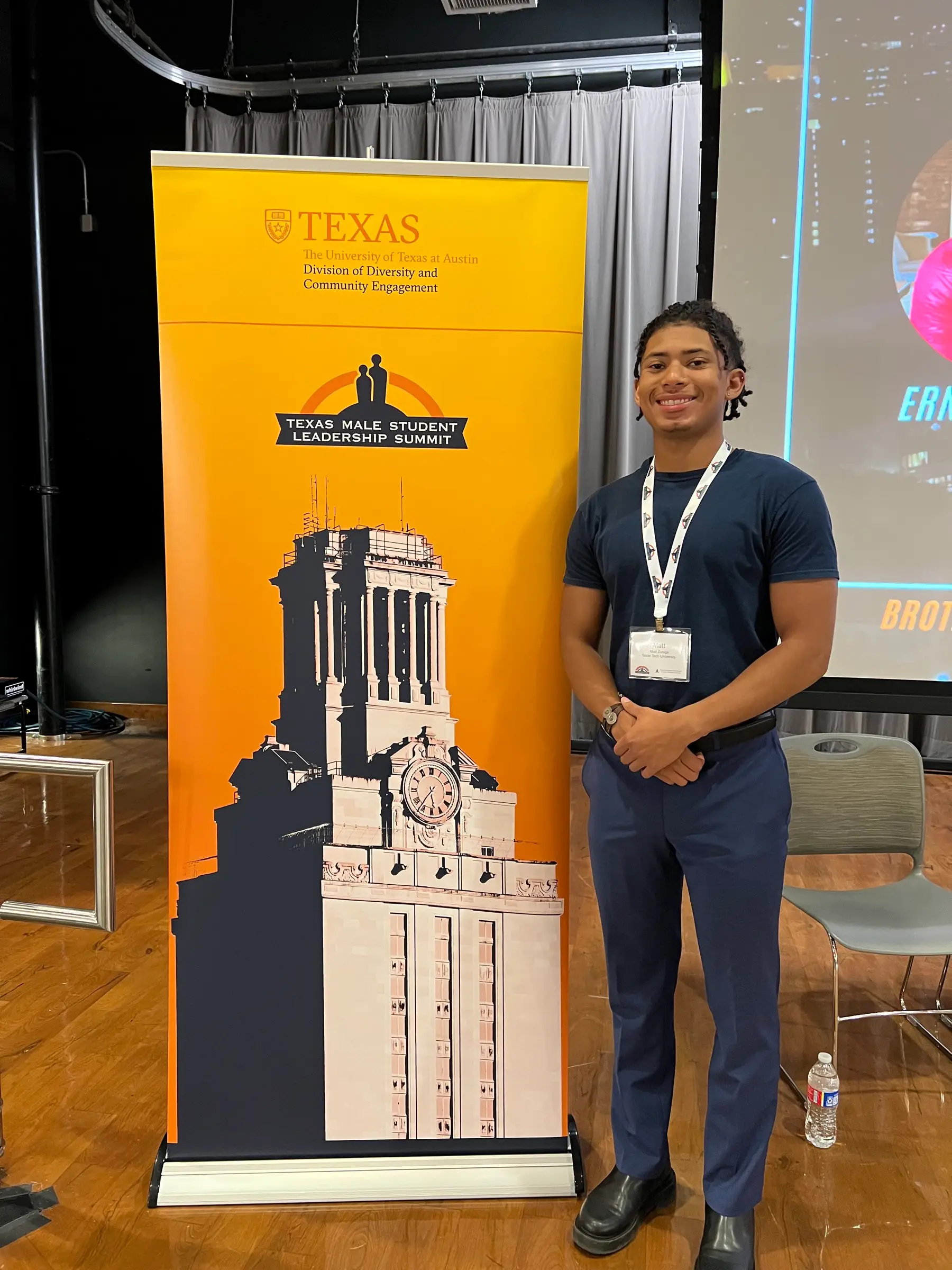 Young man with name badge beside a Texas Male Student Leadership Summit banner showing the UT Tower
