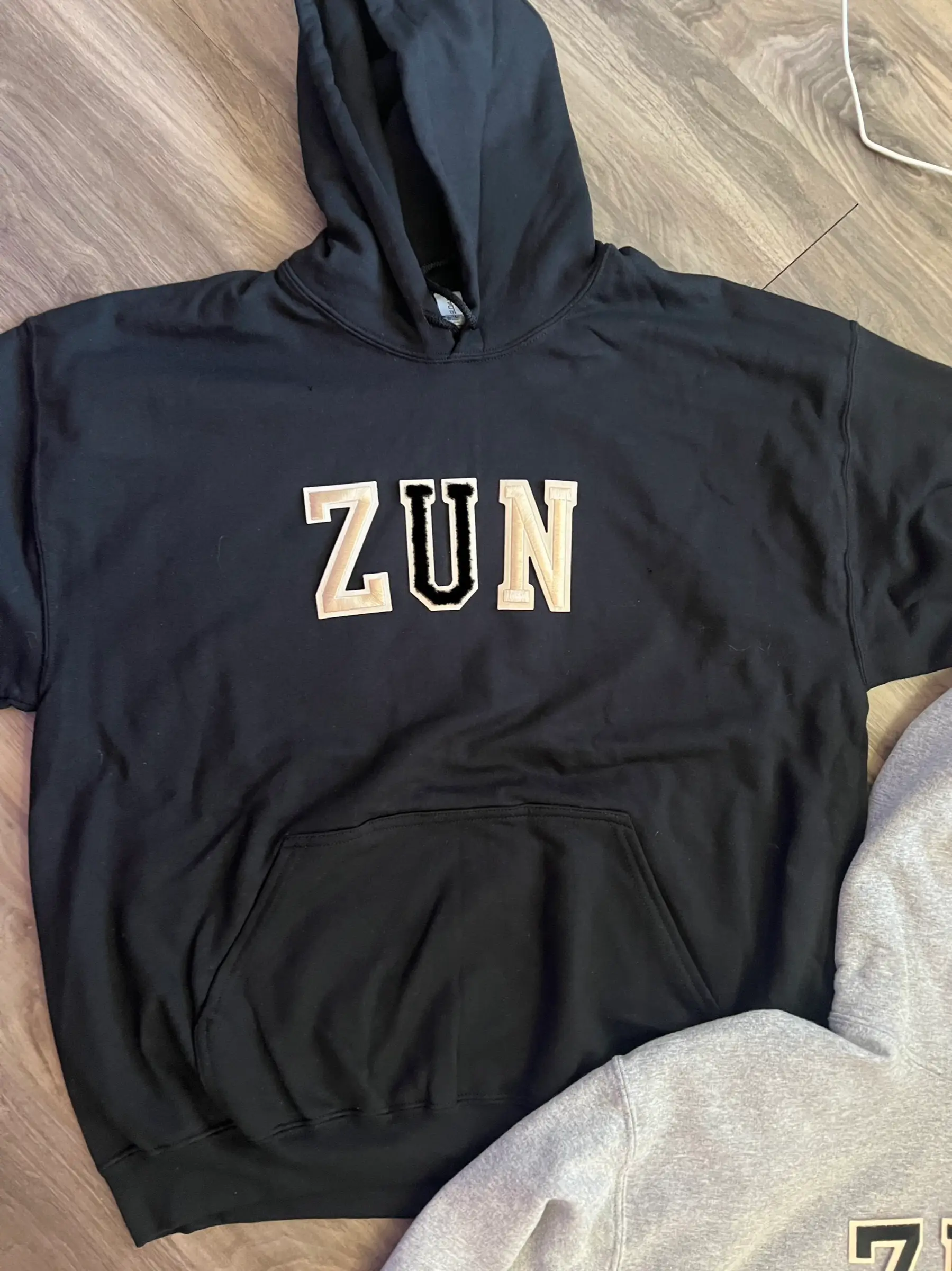 Black hoodie on wood floor with ZUN varsity letter patches, cream Z and N and a fuzzy black U
