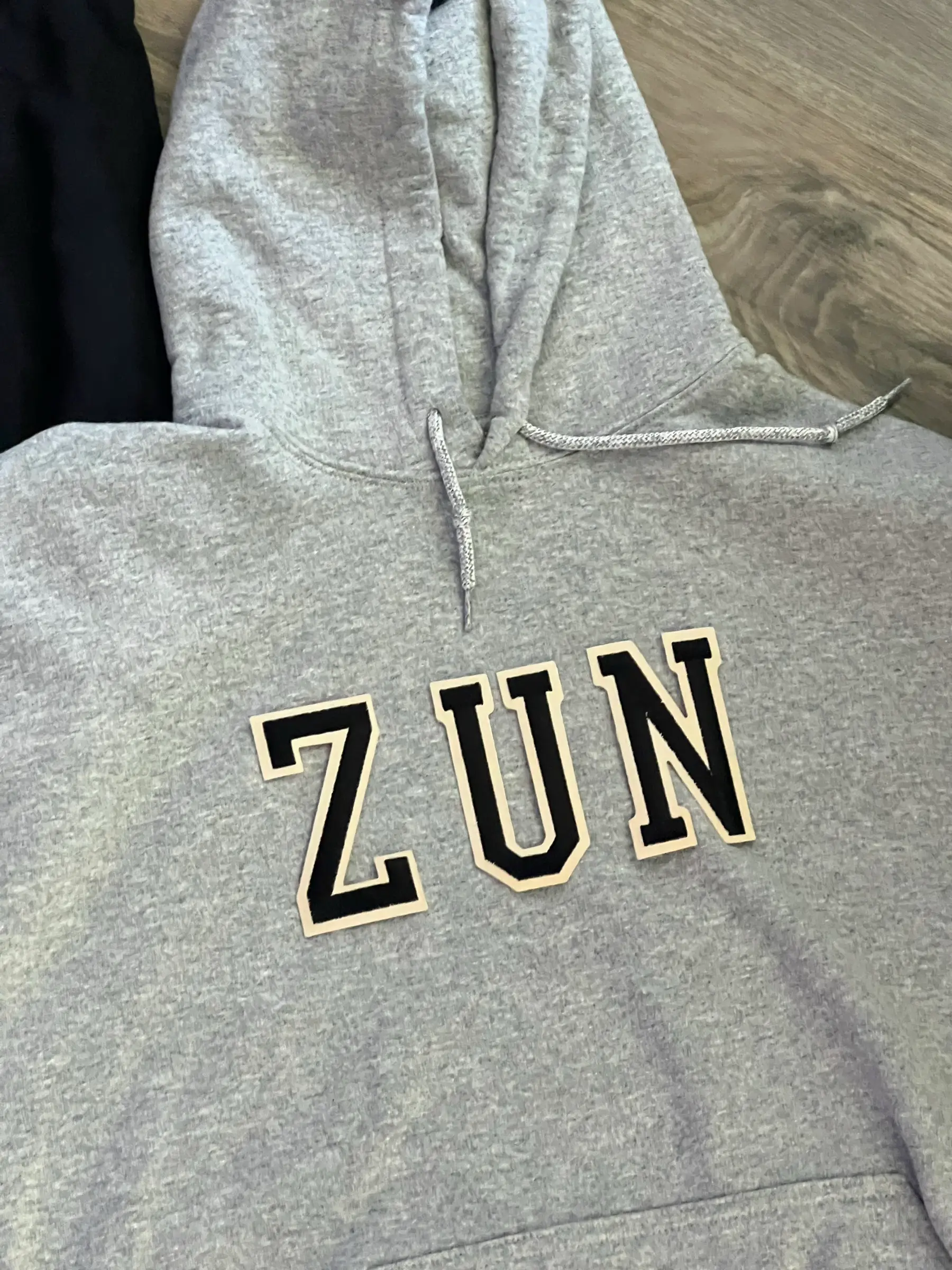 Grey hoodie close-up with black varsity ZUN letter patches outlined in cream